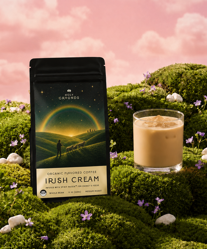 A bag of Holy Grounds Irish Cream coffee stands on a mossy hillside next to a glass of iced latte under a pink sky with clouds. The package shows a landscape illustration with a rainbow and a person silhouette.